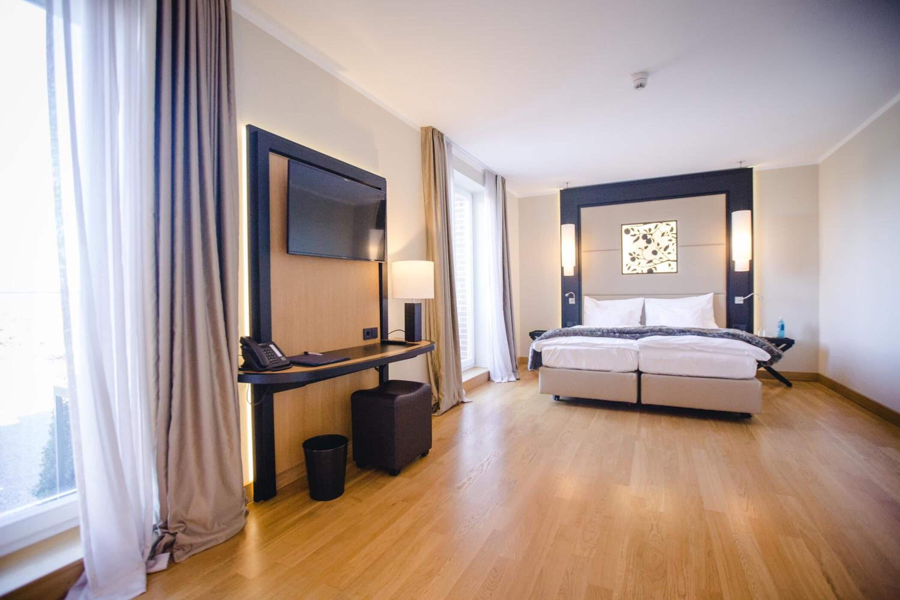 Una habitación de hotel moderna con cama, mesita de noche, lámpara y un ventanal con cortinas.