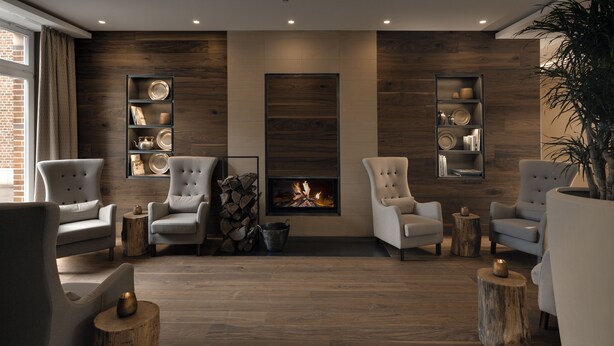 A modern living room with a fireplace, wooden walls, and a brick fireplace. There are two armchairs, a coffee table, and a side table with a candle. - Hofhotel Grothues-Potthoff (Senden)