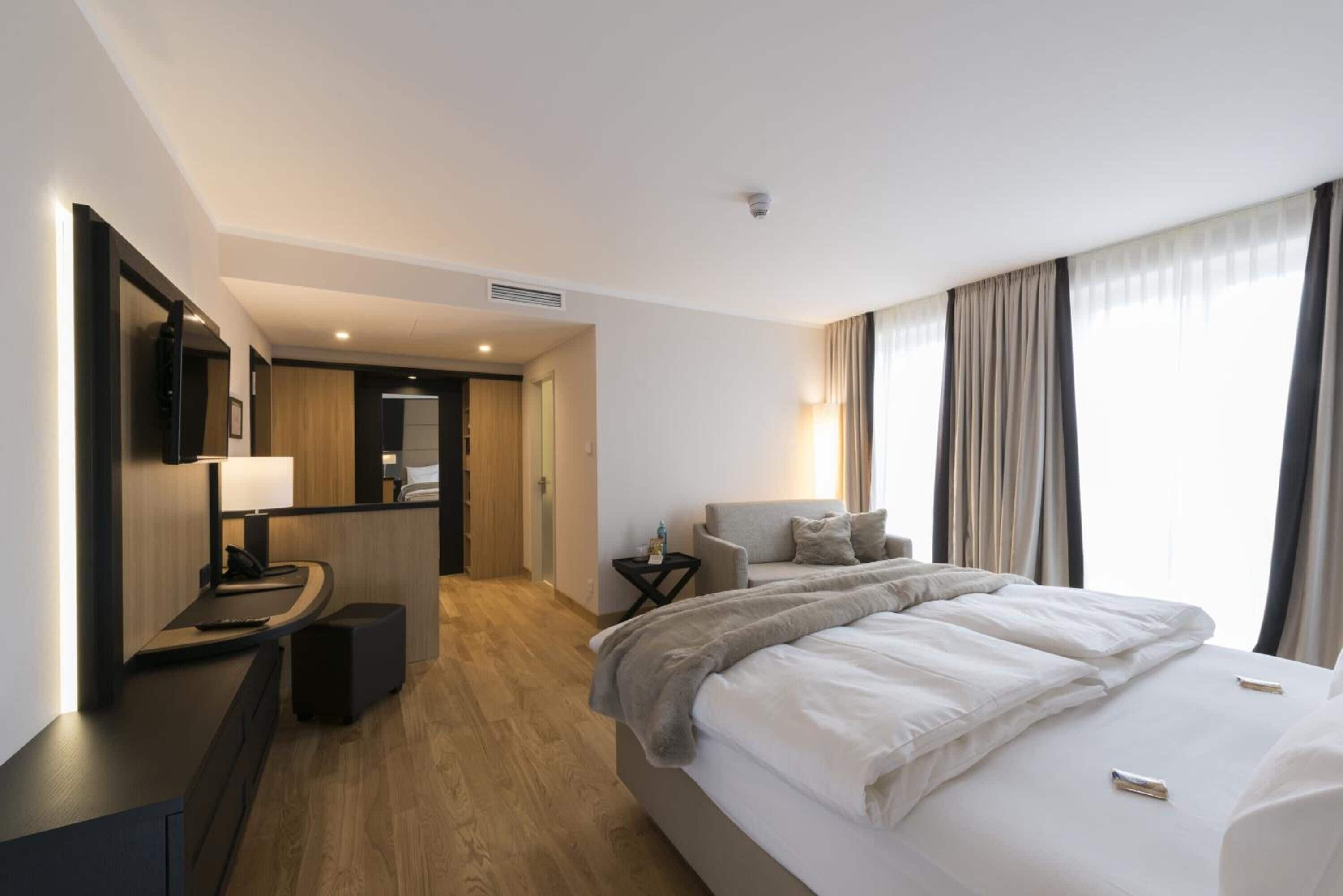 A modern hotel room with a large bed, a desk with a lamp, a small table, and a view of the city through large windows.