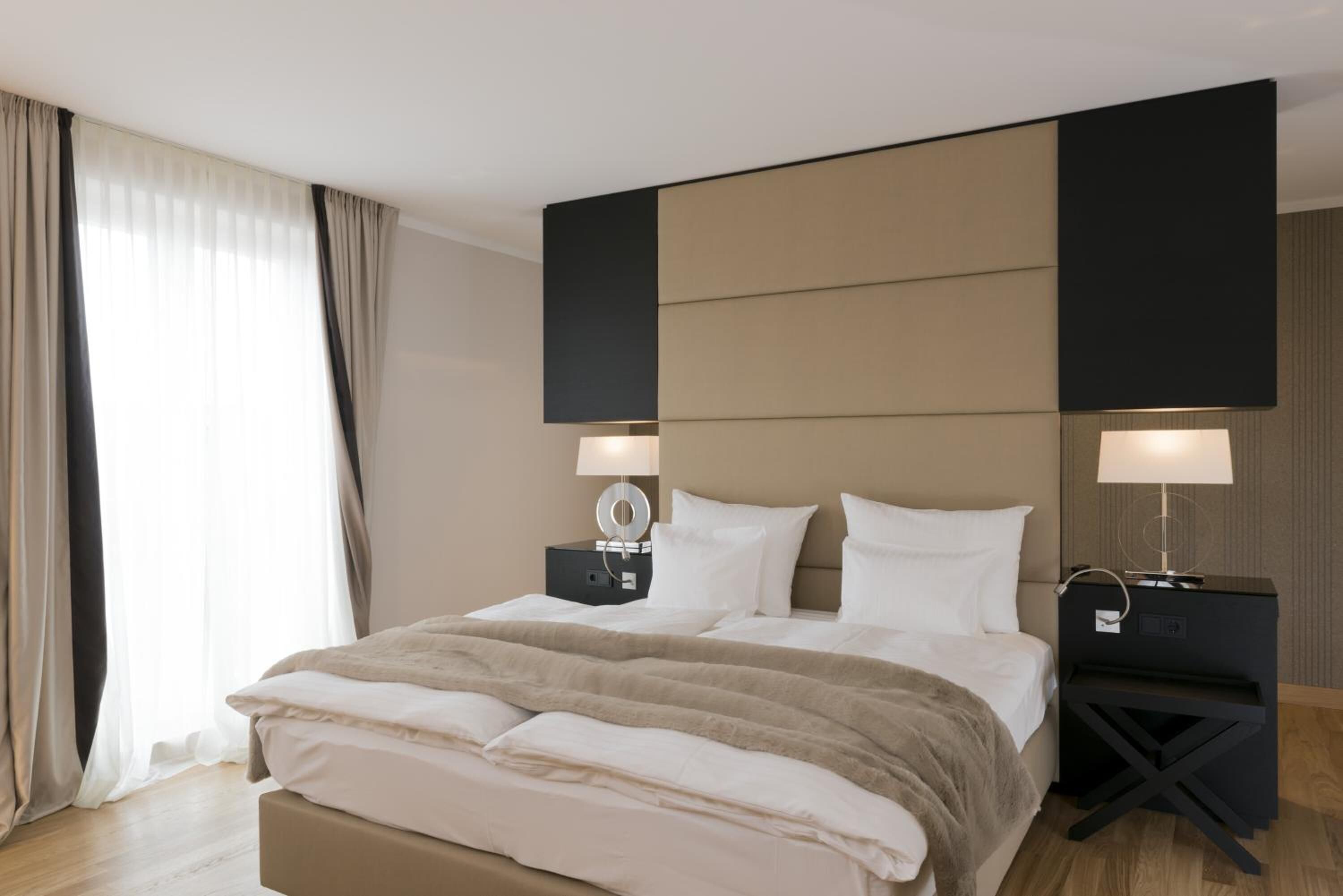 A modern hotel room with a large bed, two bedside tables with lamps, and a view of the outside through sheer curtains.