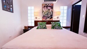 A bedroom with a large bed, two pillows with a tropical pattern, a wall-mounted air conditioner, and a window with a view.