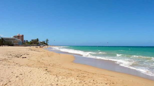 A sandy beach with waves, a clear sky, and buildings in the background.