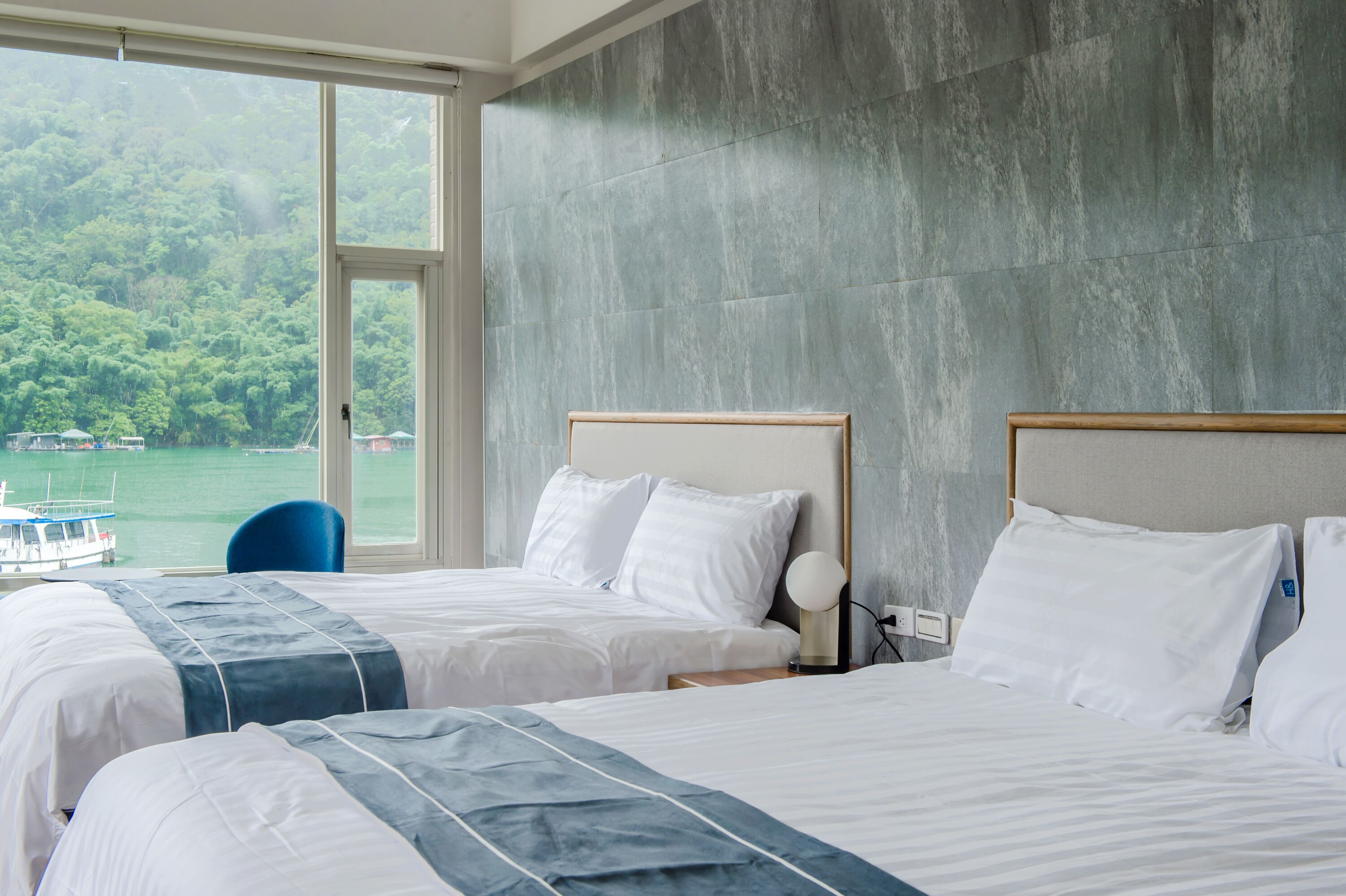 A hotel room with two beds, a large window with a view of a lake and boats, and a stone-textured wall.