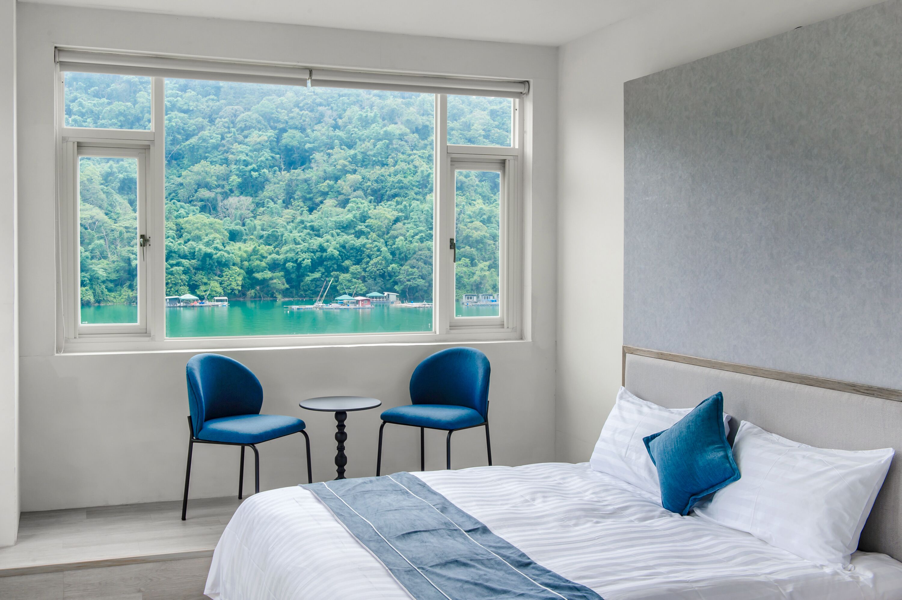 Une chambre avec une grande fenêtre, deux chaises, une petite table et un lit avec une literie blanche et un oreiller bleu.