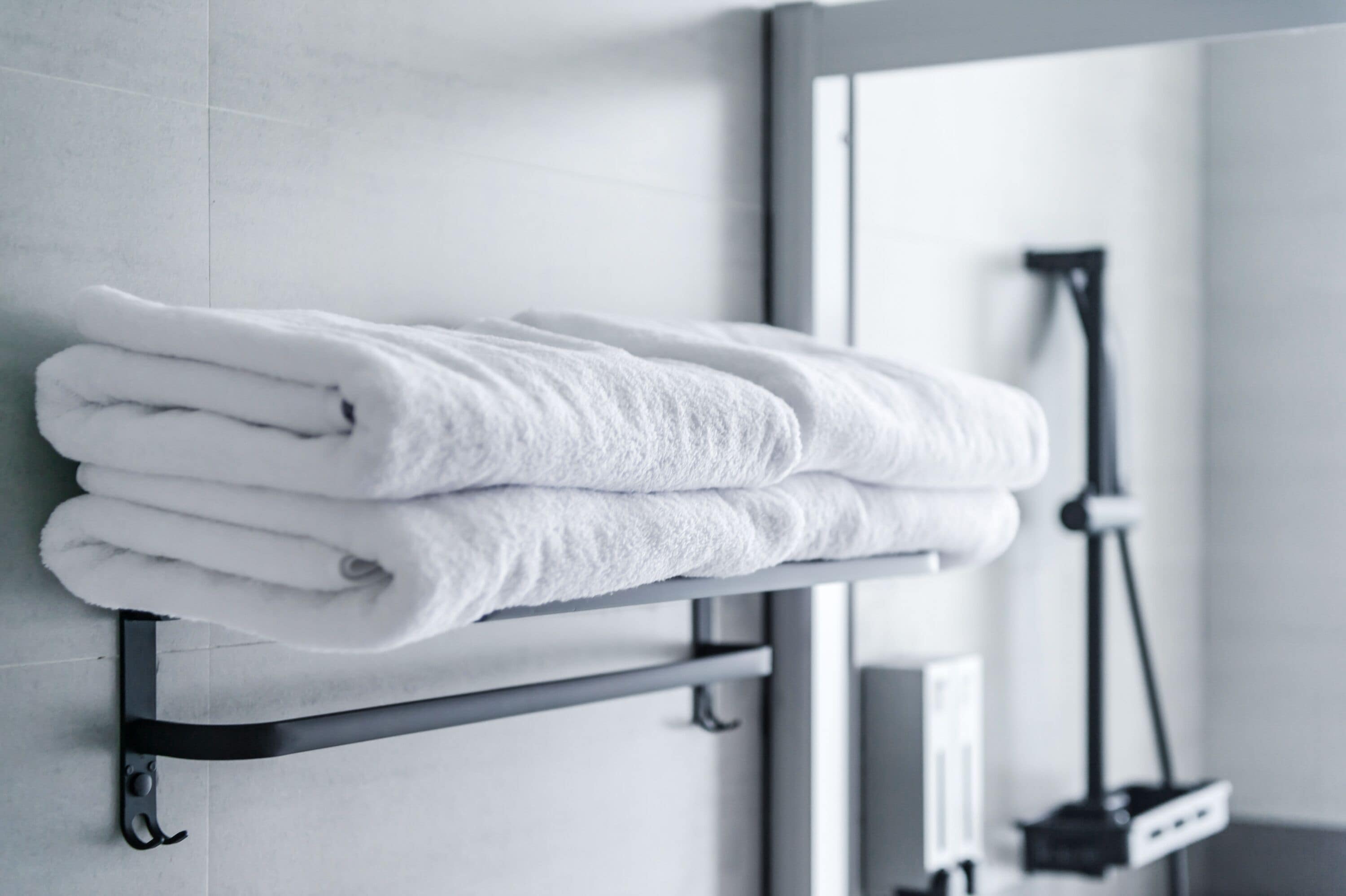 A neatly folded white towel stacked on a metal towel rack in a bathroom with a shower head visible in the background.