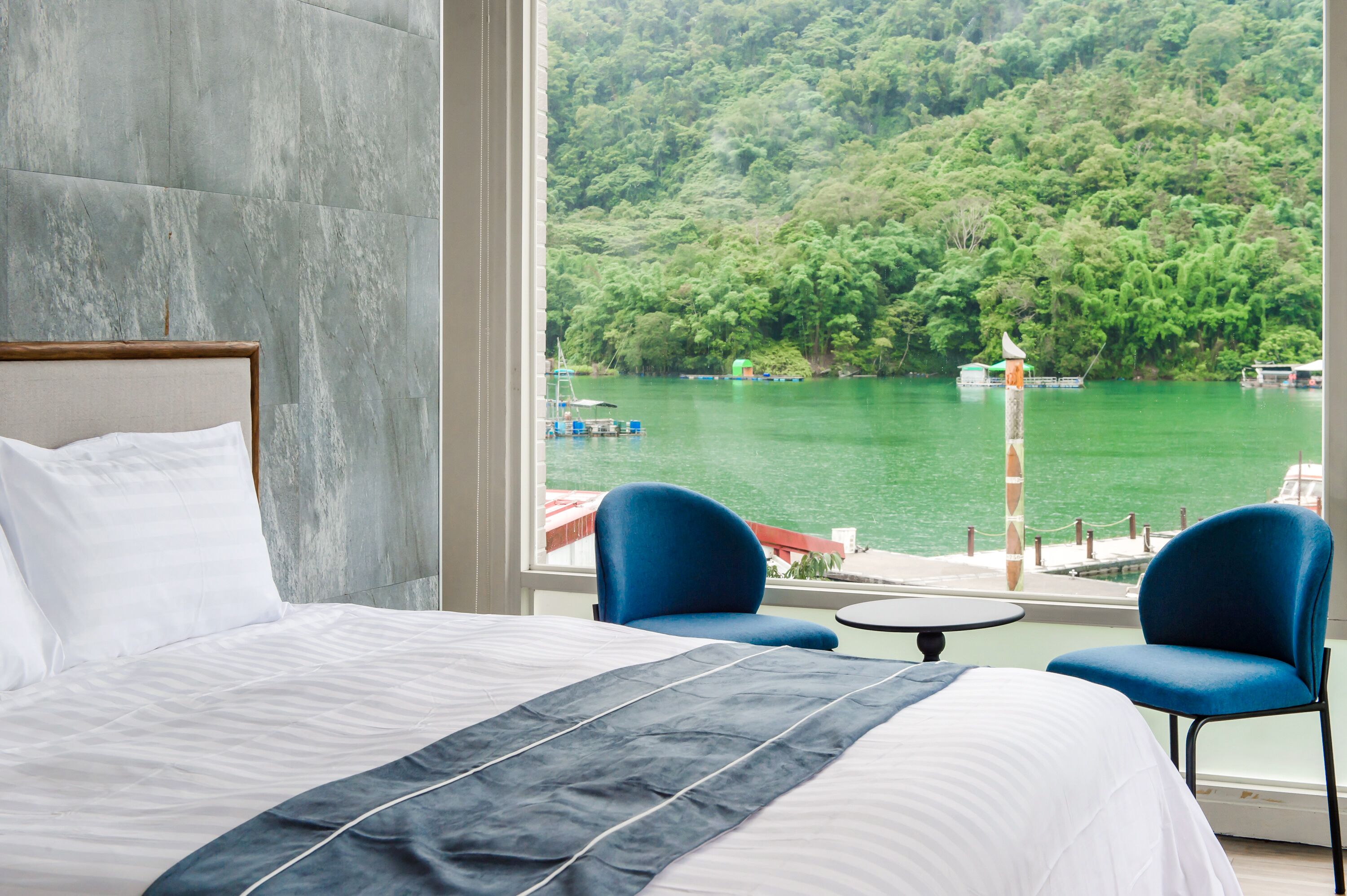 A hotel room with a large bed, two blue chairs, and a view of a lake with boats.