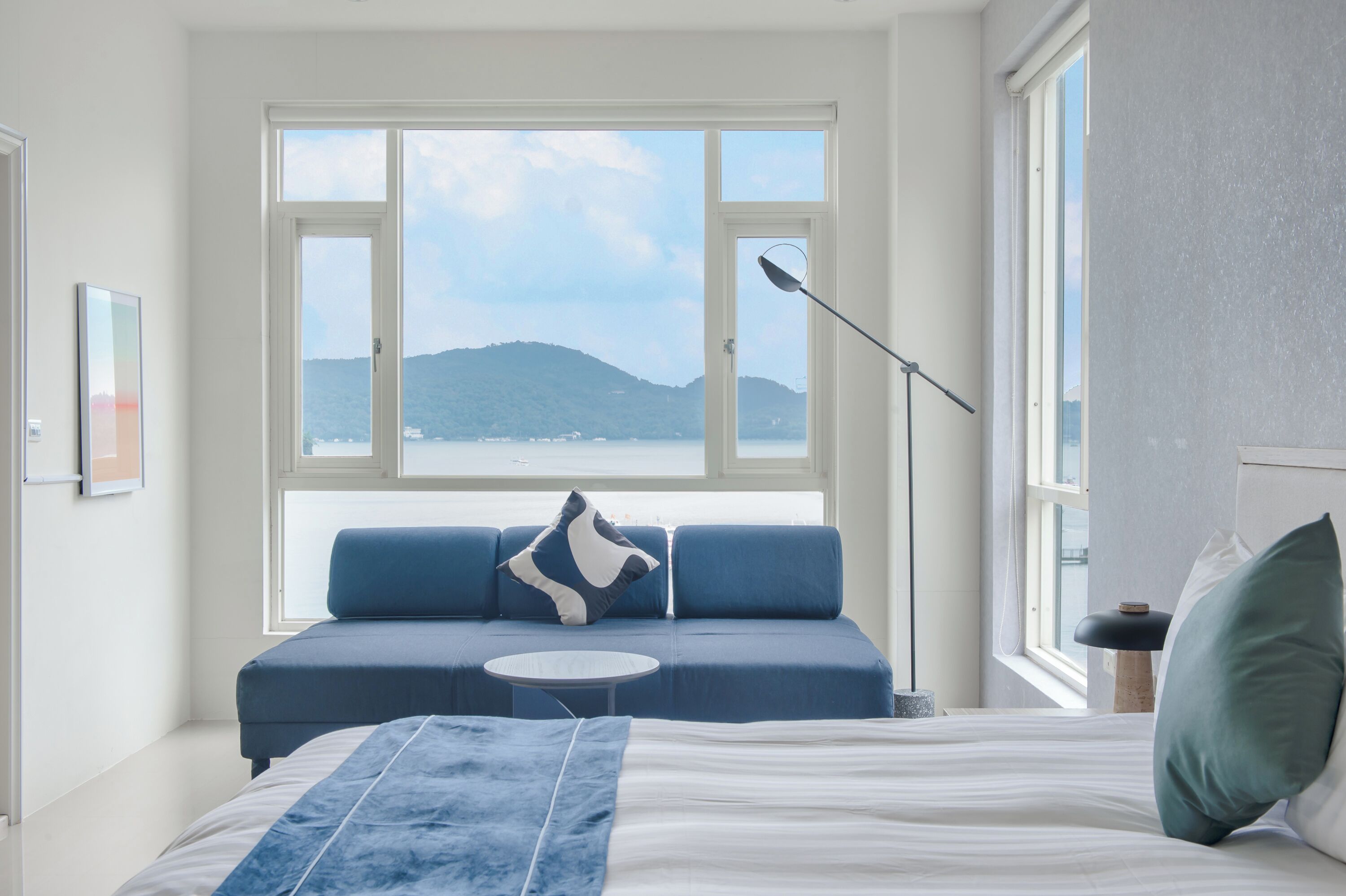 Een slaapkamer met een groot raam, een bed, een blauwe bank en een klein, rond tafeltje.