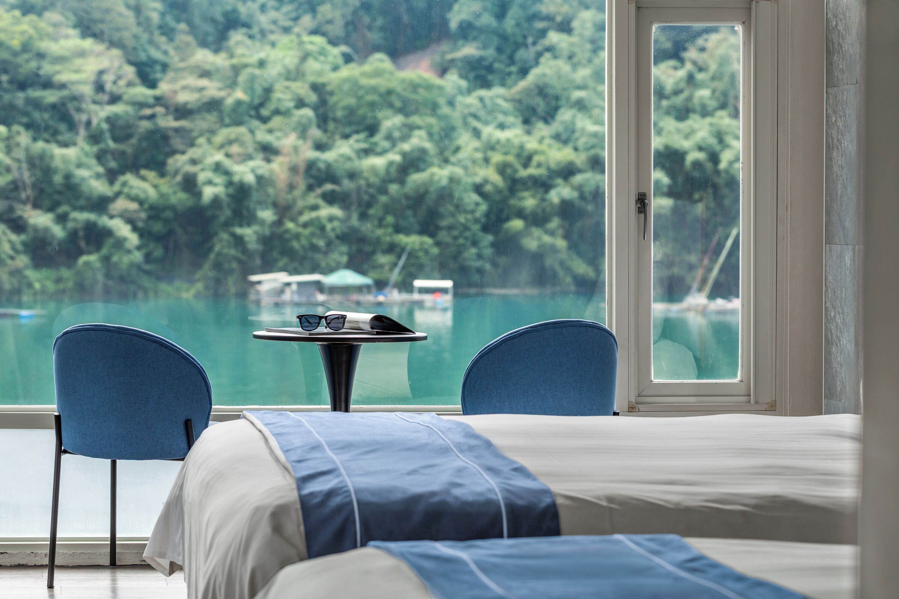 A room with a bed, two chairs, a small table with glasses, and a view of a lake and forest.
