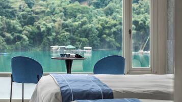 A room with a bed, two chairs, a small table with glasses, and a view of a lake and forest.