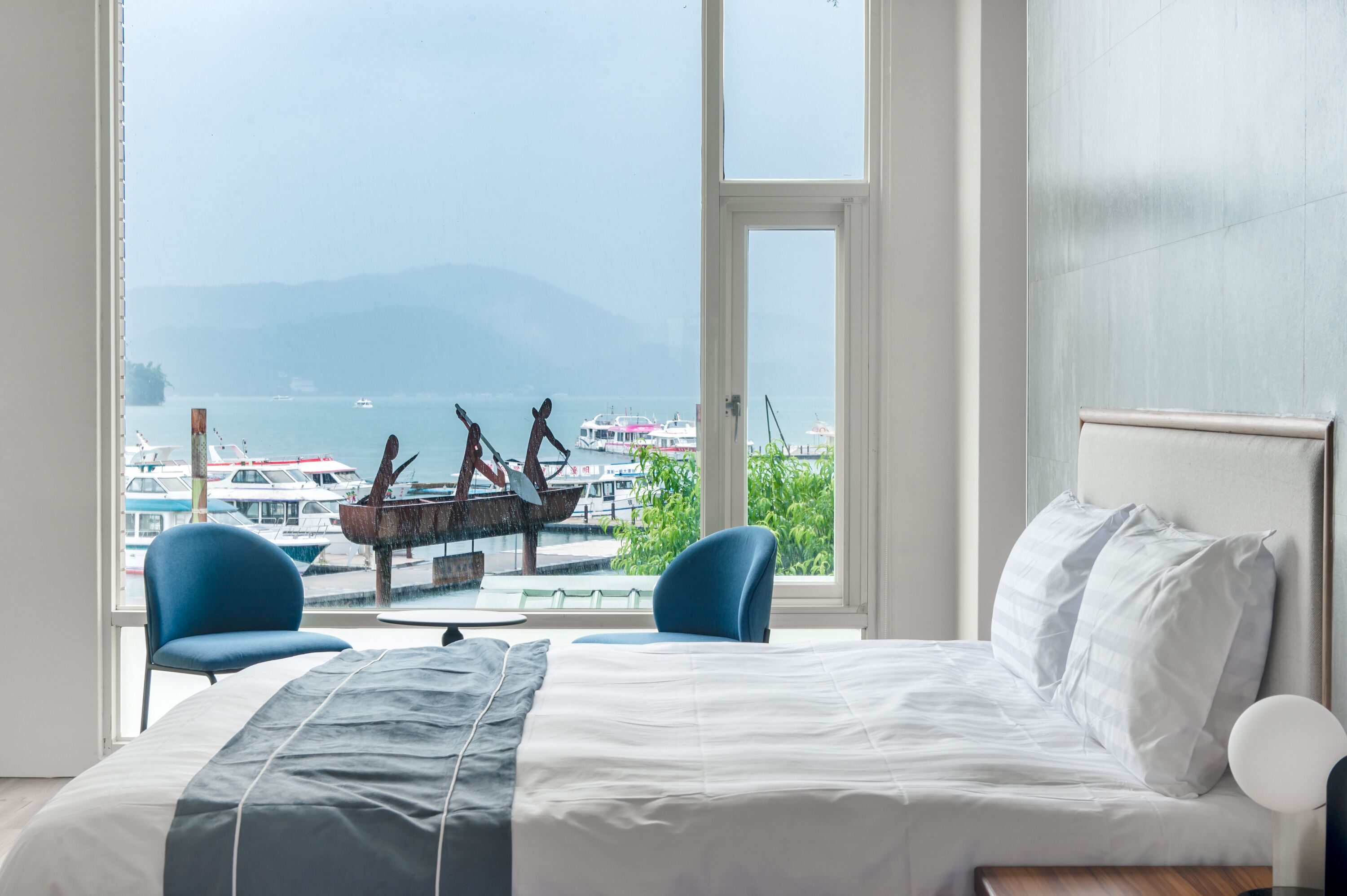 Habitación de hotel con una cama grande, dos sillas azules y vistas a barcos y montañas a través de un ventanal amplio.
