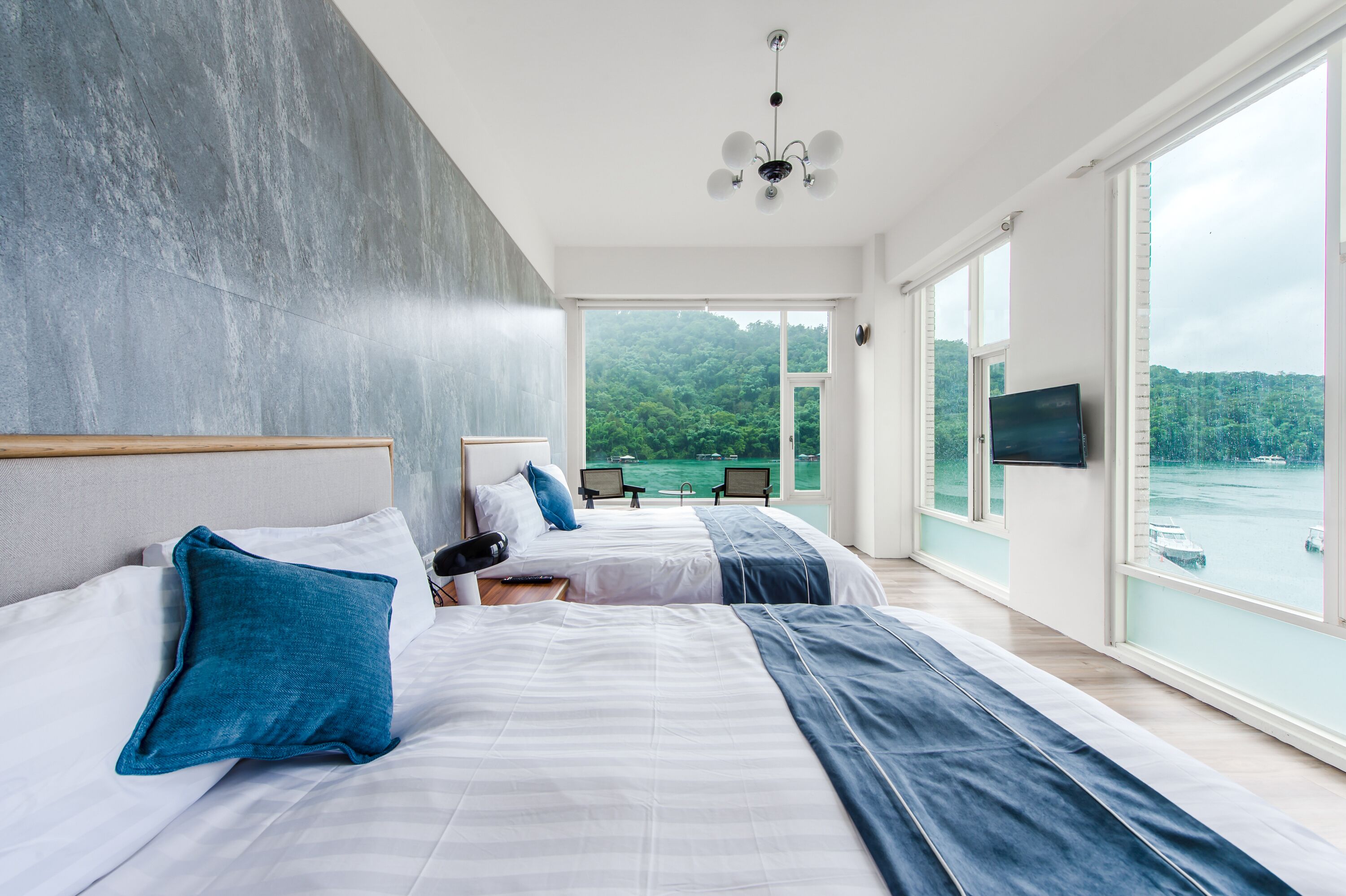 A modern hotel room with two beds, a large window with a view of a lake, and a flat-screen TV mounted on the wall.