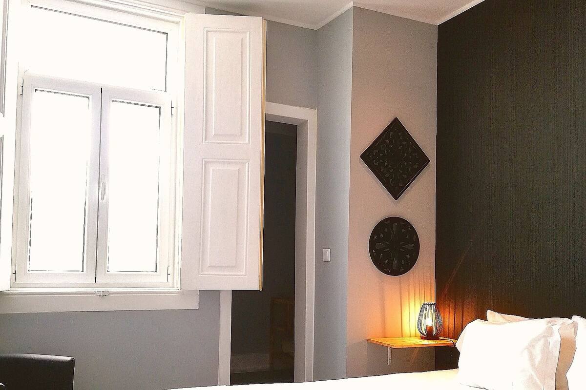 A bedroom with a large bed, a desk, and a chair. There is a window with white shutters, a wall clock, and a bedside table with a lamp.