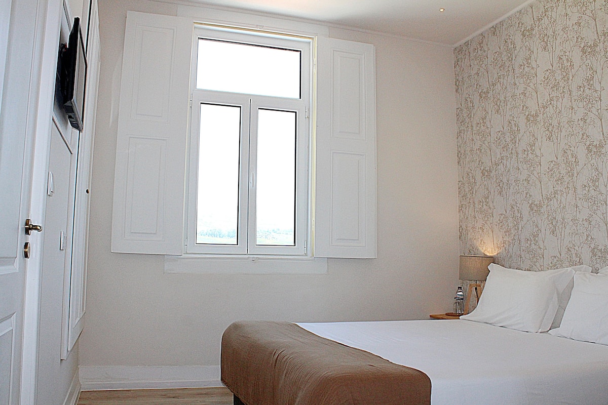 A bedroom with a bed, a window with shutters, a nightstand, and a wall with floral wallpaper.