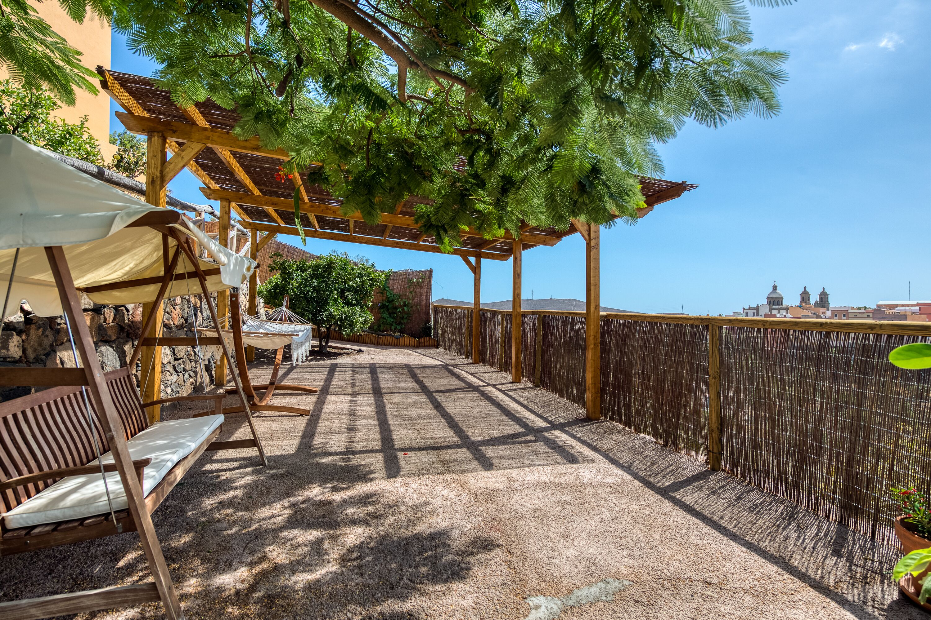 A covered outdoor seating area with wooden furniture and a pergola providing shade, overlooking a cityscape.