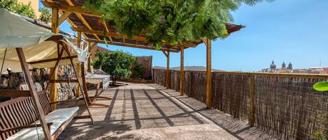 A covered outdoor seating area with wooden furniture and a pergola providing shade, overlooking a cityscape.