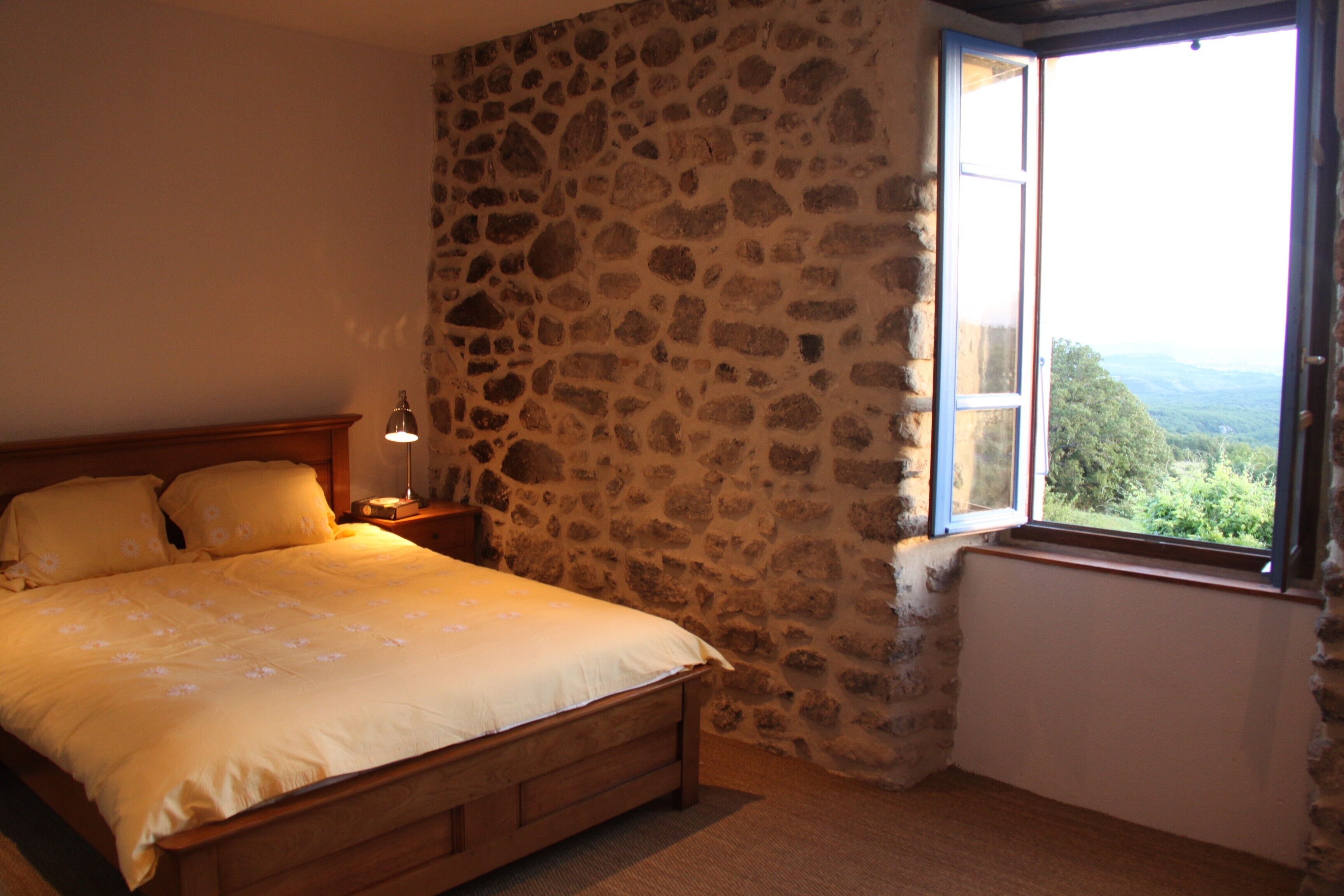 A bedroom with a stone wall, a wooden bed, and a window with a view of the countryside.