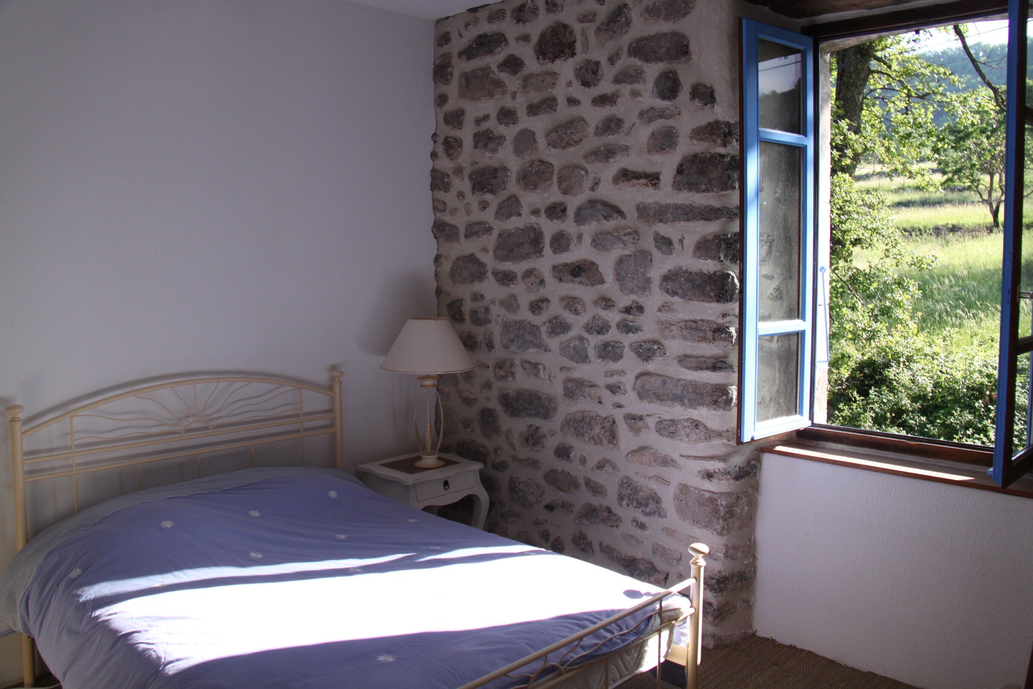 A bedroom with a stone wall, a bed with a blue bedspread, a nightstand with a lamp, and a window with a view of greenery.