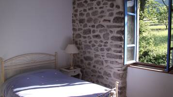 A bedroom with a stone wall, a bed with a blue bedspread, a nightstand with a lamp, and a window with a view of greenery.