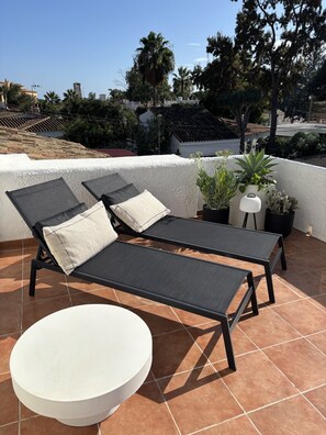 A rooftop terrace with two lounge chairs and a round table, surrounded by potted plants.