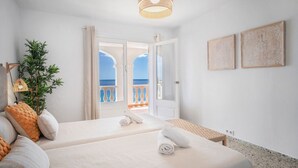 Habitación de hotel con dos camas, un balcón con vistas al mar, una lámpara colgante y dos cuadros enmarcados en la pared.