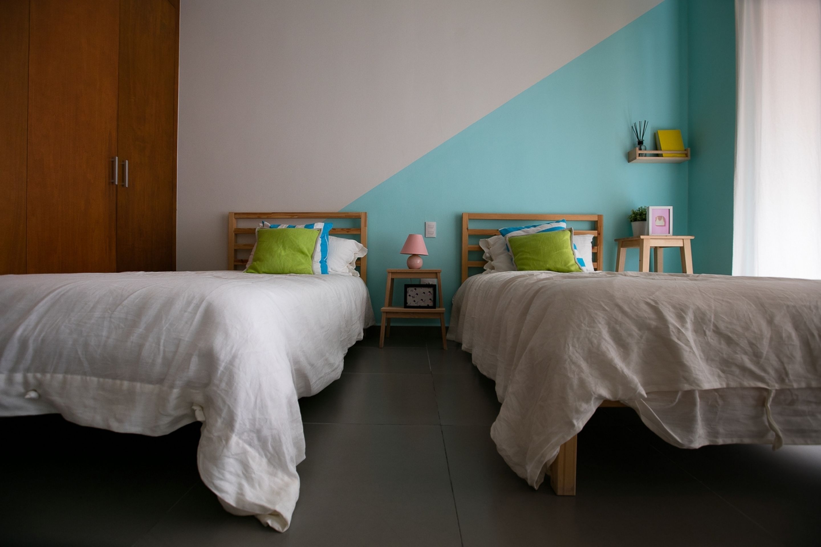 A bedroom with two beds, a wooden wardrobe, a nightstand with a lamp, and a shelf with decorative items.