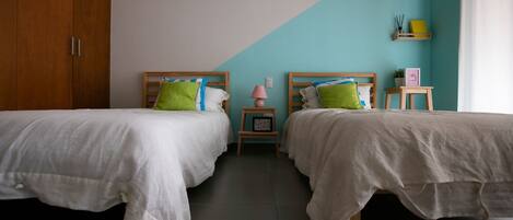 A bedroom with two beds, a wooden wardrobe, a nightstand with a lamp, and a shelf with decorative items.
