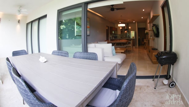A modern dining area with a large table and wicker chairs, a sofa, and a kitchen visible through an open doorway. - Fabulous 3BR Condo steps away from 5th Avenue and beach by Happy Adddress (Playa del Carmen)