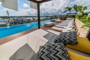 Un área de piscina en la azotea con sillones y cojines, una terraza de madera y vistas a la ciudad.