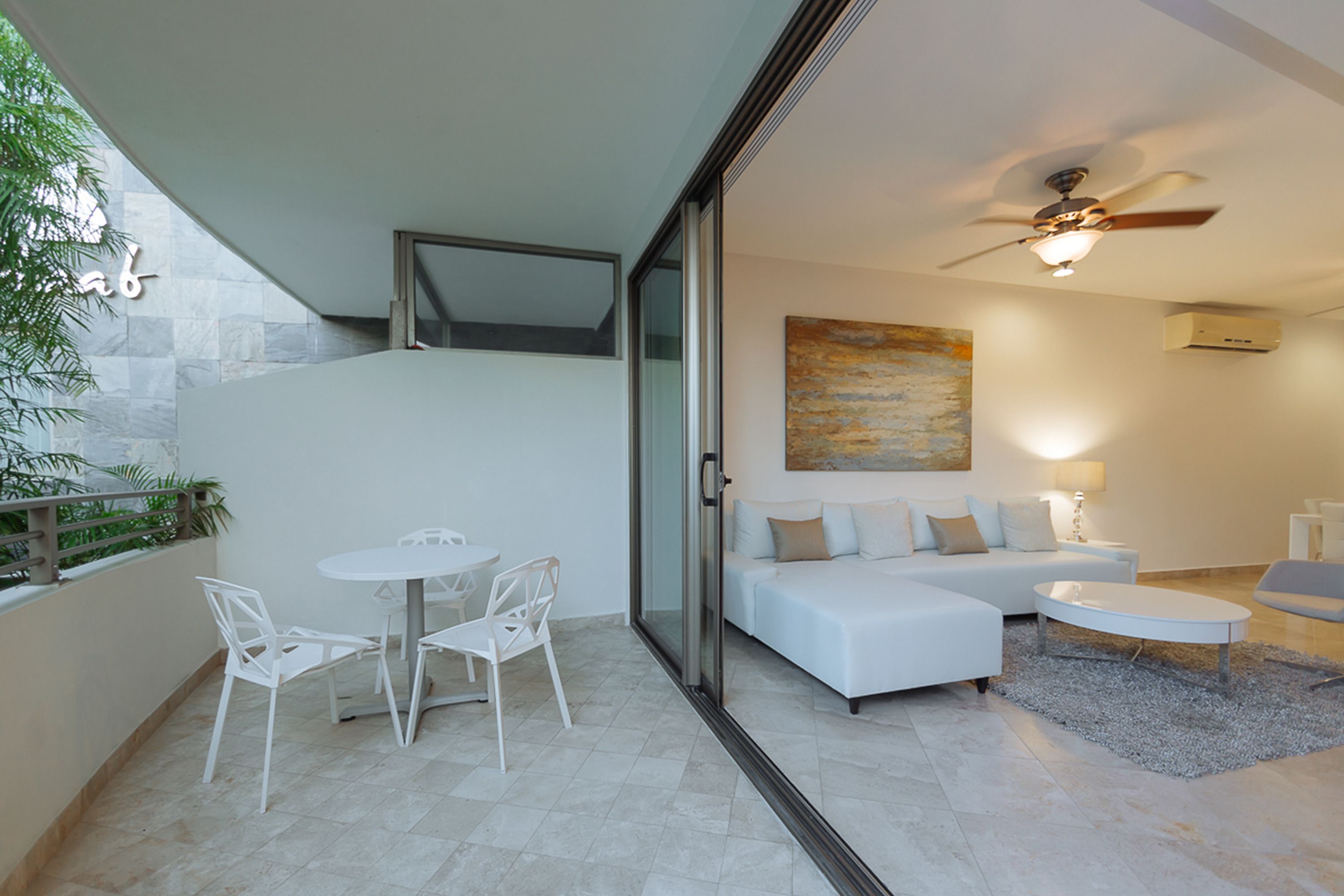 A modern living room with a sofa, coffee table, and chairs. There is a ceiling fan, a painting on the wall, and a glass door leading to an outdoor area with a table and chairs.