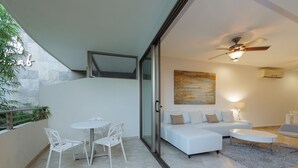 Un soggiorno moderno con divano, tavolino e poltrone. Si nota una ventola a soffitto, un quadro appeso al muro e una porta vetrata che dà su una zona esterna con tavolo e sedie.