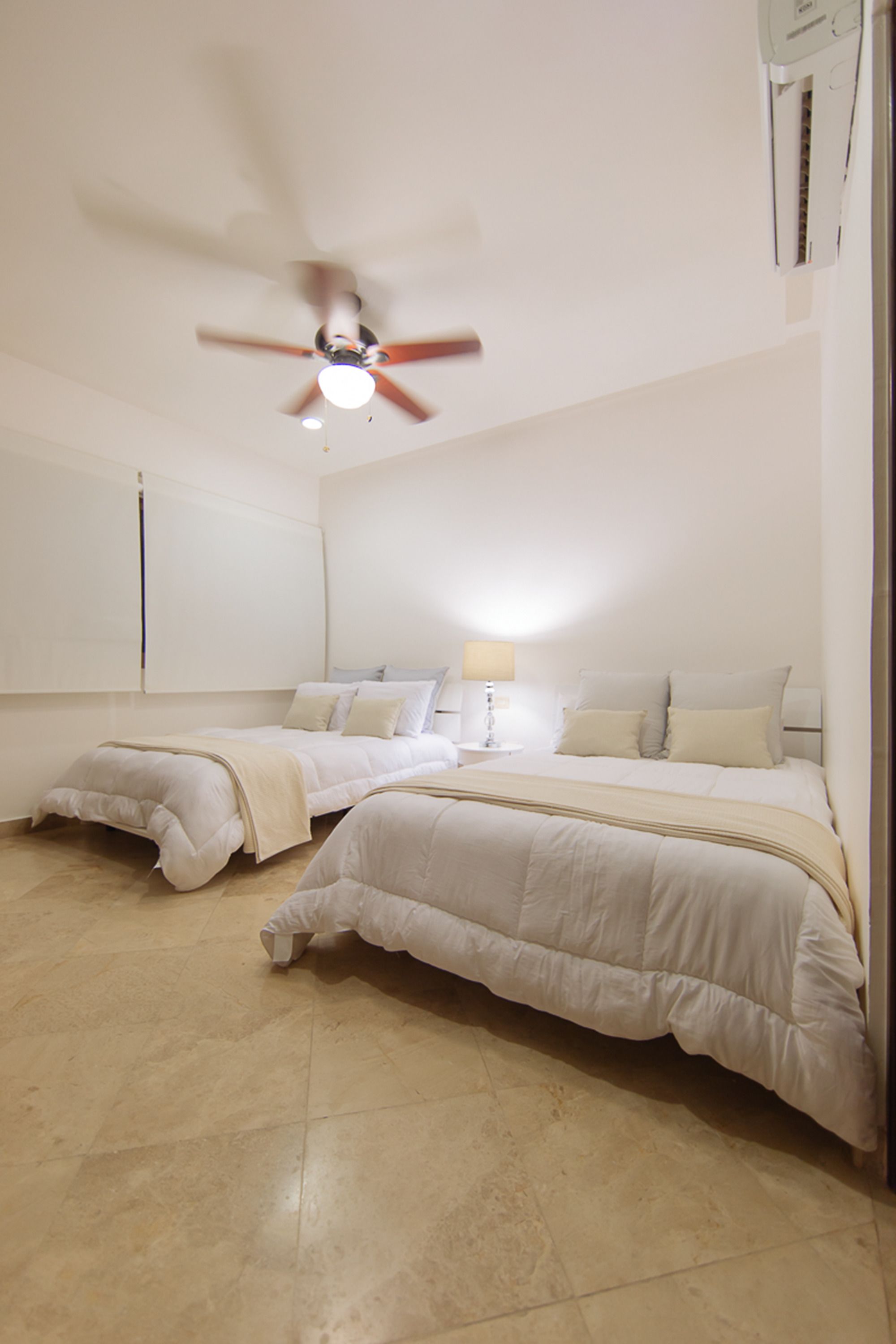 A hotel room with two beds, a ceiling fan, a nightstand with a lamp, and a wall-mounted air conditioning unit.
