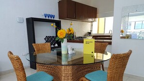 Un comedor con una mesa redonda de vidrio, sillas de mimbre con cojines azules y un jarrón con flores amarillas.