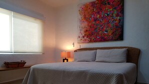 Un dormitorio con cama, mesita de noche, lámpara, ventana y un cuadro abstracto colorido en la pared.