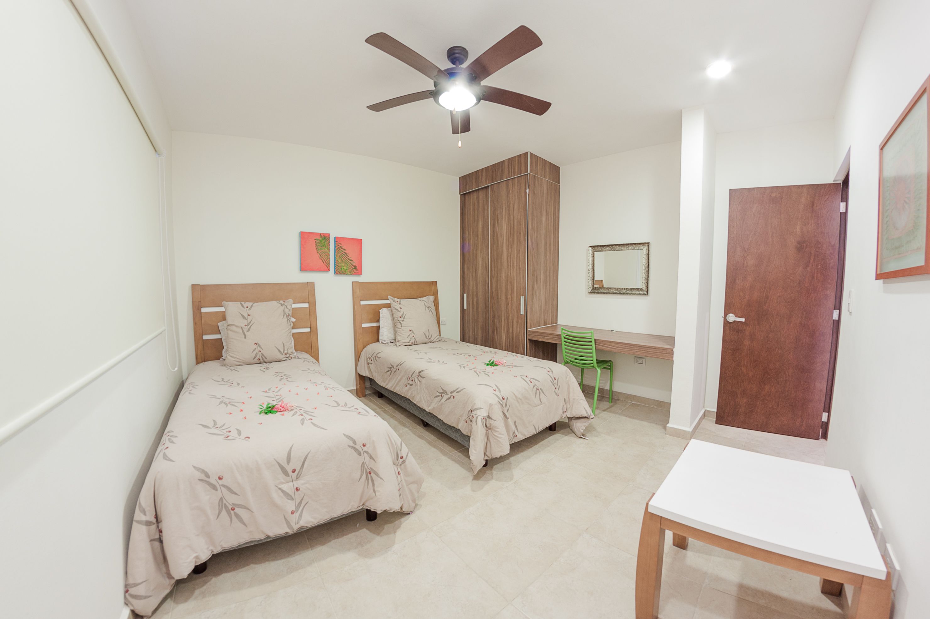 Quarto com duas camas, uma escrivaninha, uma cadeira, um ventilador de teto e uma porta.