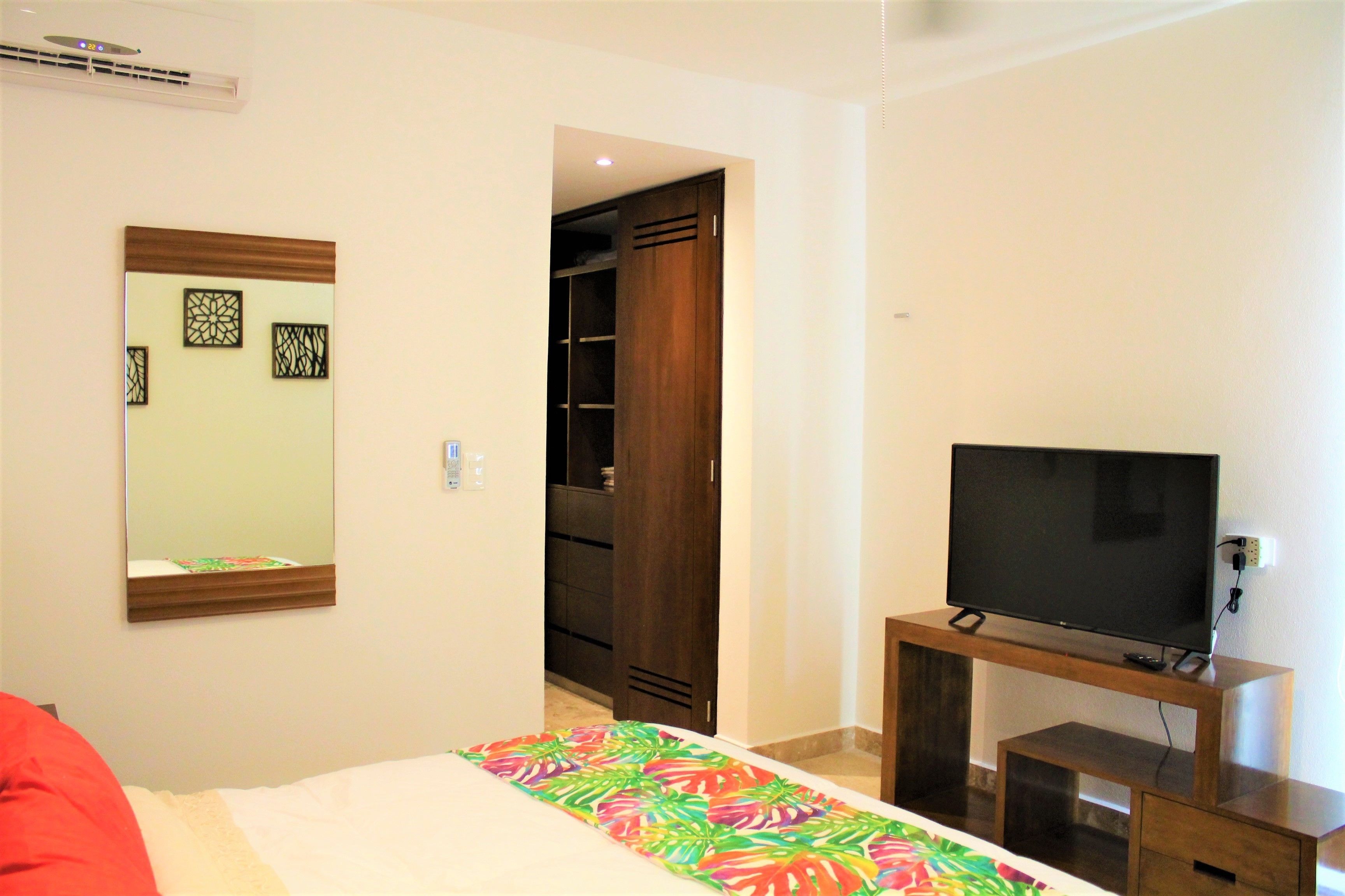 A bedroom with a bed, a TV on a wooden stand, a wardrobe, and a mirror on the wall.