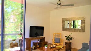 Sala de estar con televisor, muebles de madera, un sofá y un ventanal con vistas a la vegetación.