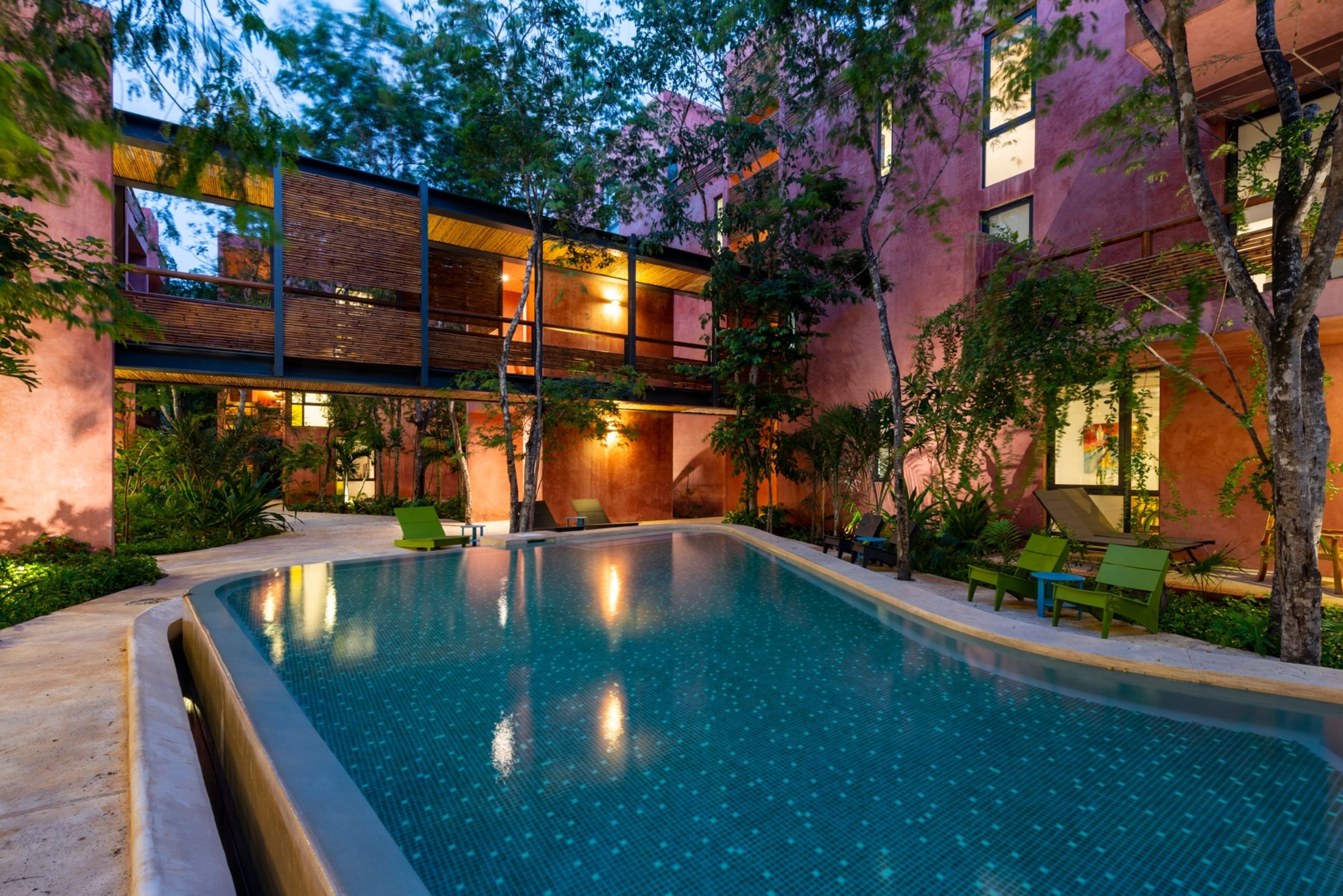 A swimming pool with a wooden deck and lounge chairs, surrounded by trees and a building with large windows.