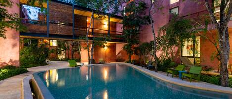 A swimming pool with a wooden deck and lounge chairs, surrounded by trees and a building with large windows.