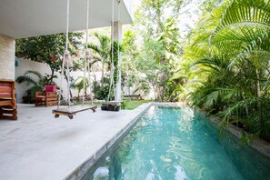 Un espace piscine agrémenté d’un fauteuil suspendu, entouré d’une végétation luxuriante et bordé d’un mur blanc.