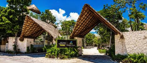A tropical resort entrance with thatched-roof structures and a sign indicating arrival at Puerto Zamá.
