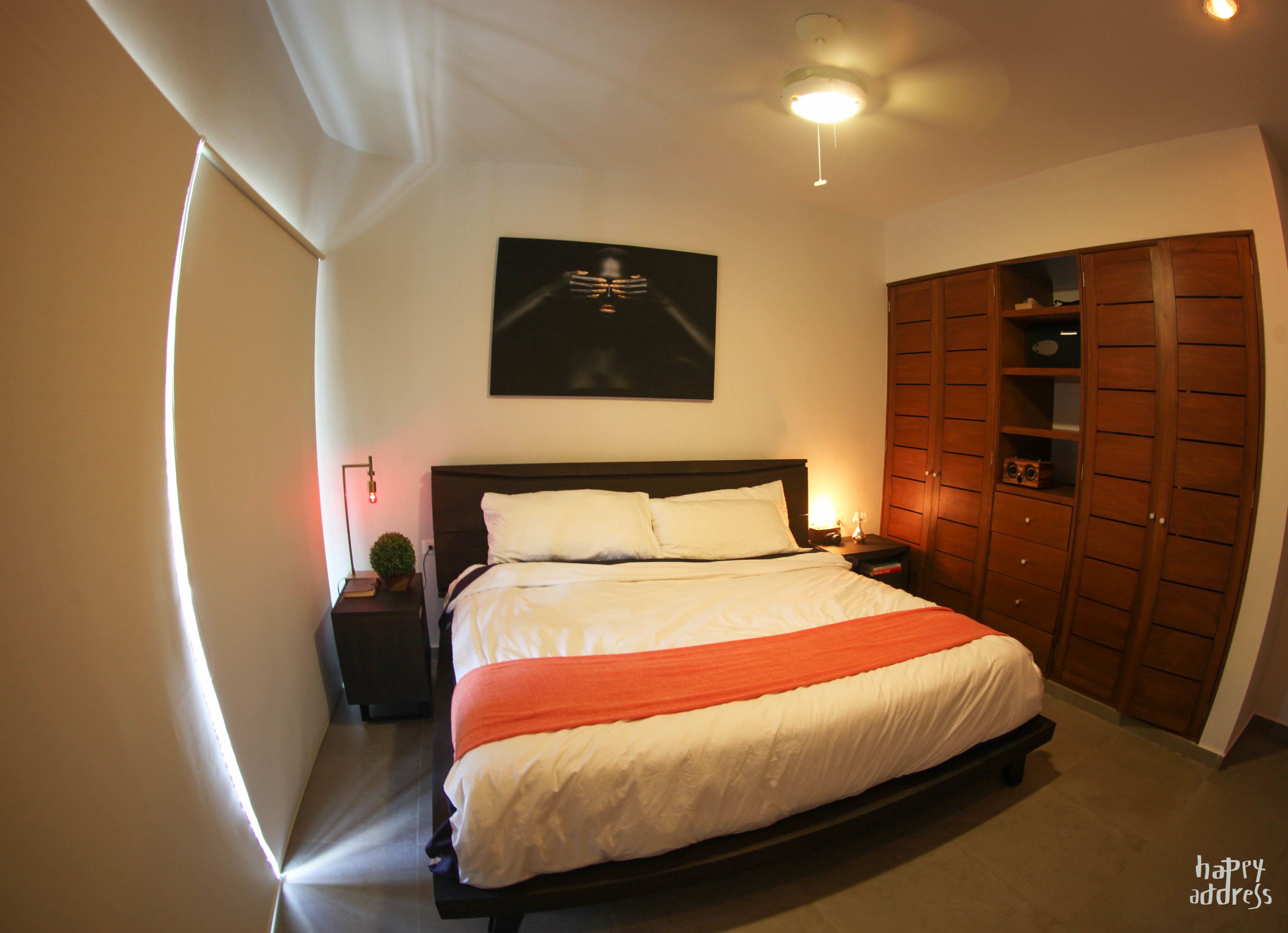 A bedroom with a large bed, a nightstand, a wall-mounted light, a ceiling fan, and a wooden wardrobe.