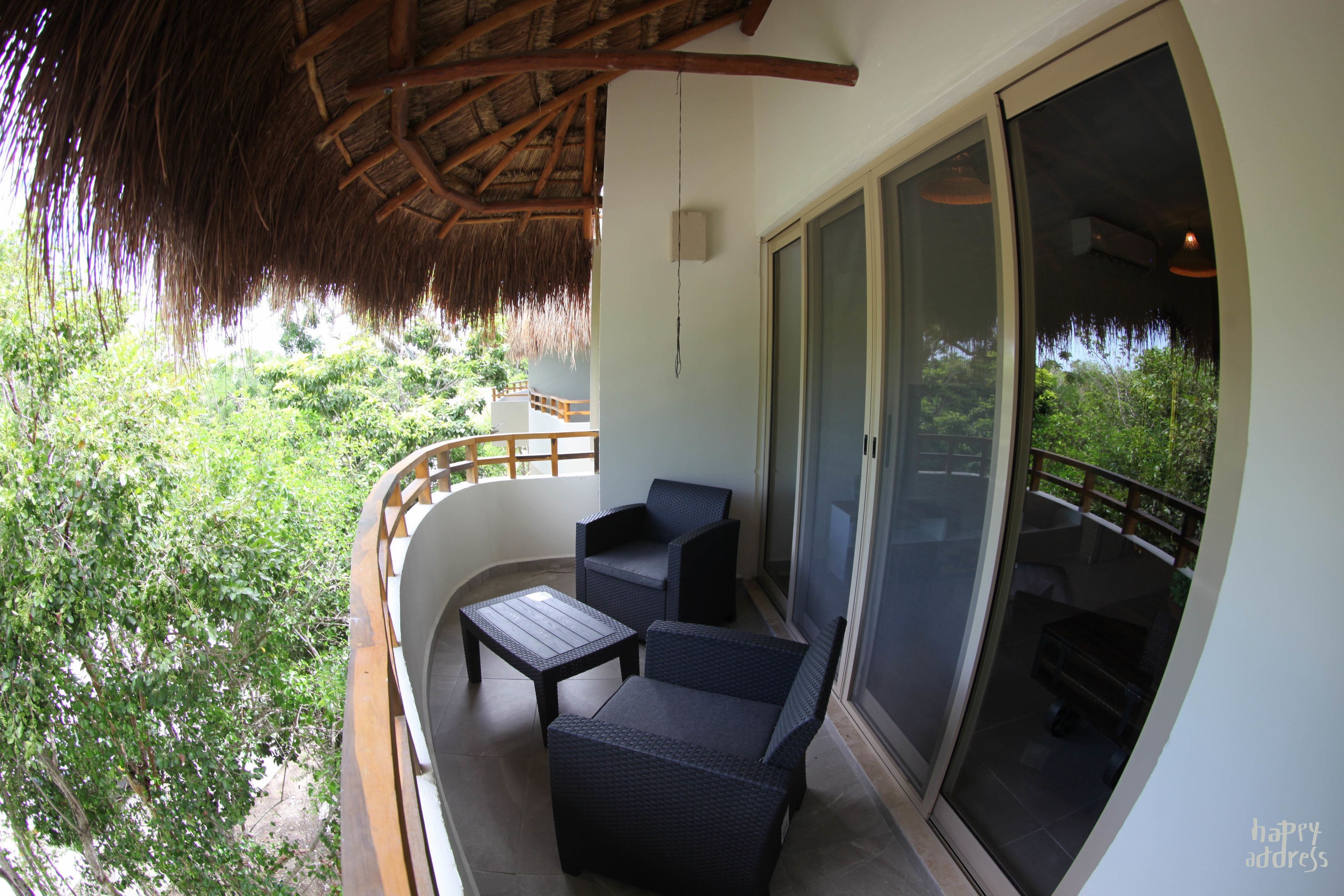 A balcony with wicker furniture, a thatched roof, and a sliding glass door leading to a lush garden.
