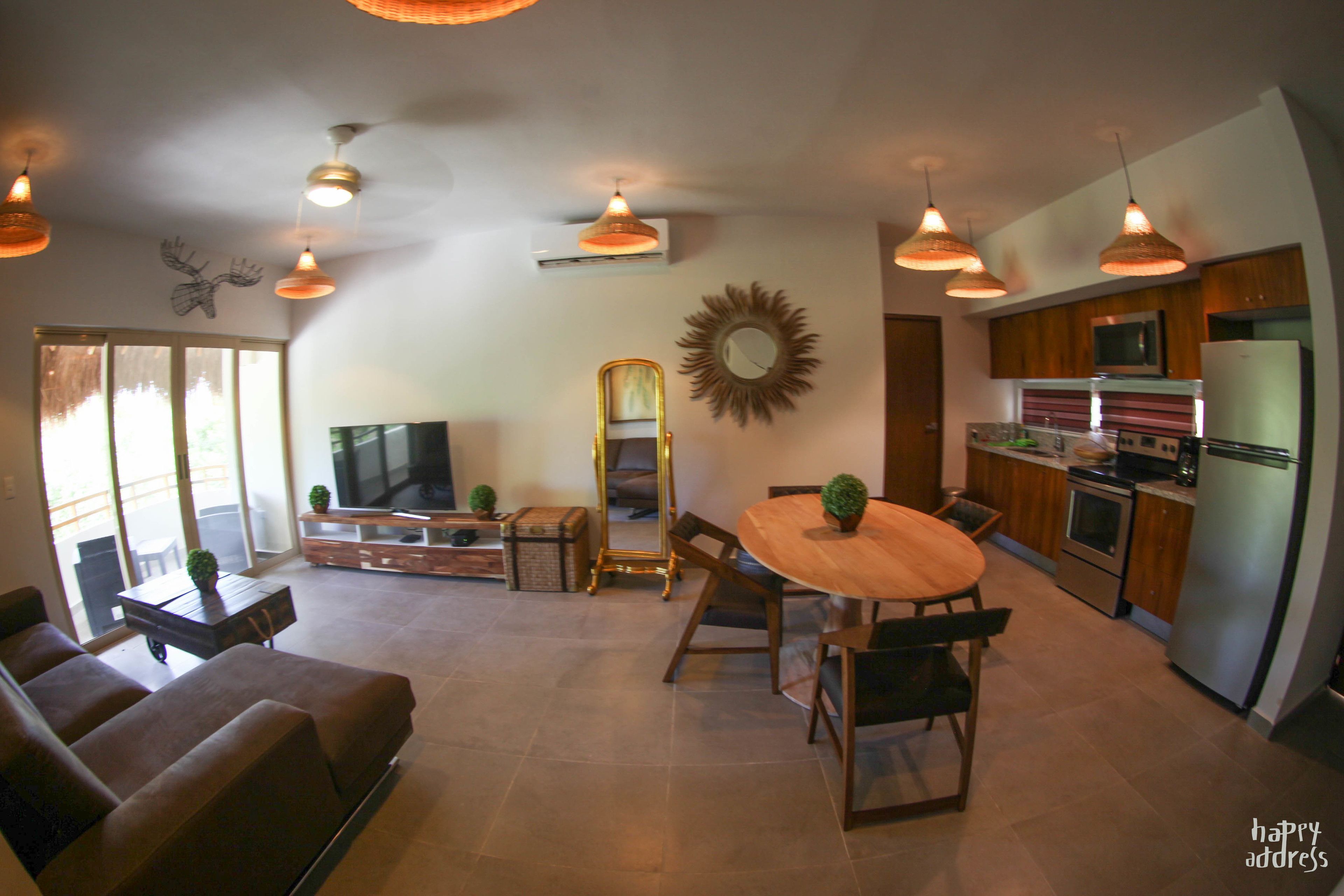 A modern living room with a sofa, dining table, and chairs. There is a television, a mirror, and a sunburst decoration on the wall. The kitchen is visible with cabinets and appliances.