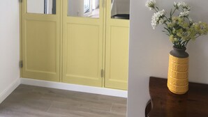 Ein Zimmer mit einem gelben Kleiderschrank, einem Holztisch mit Blumenvase und einem Bett im Hintergrund.