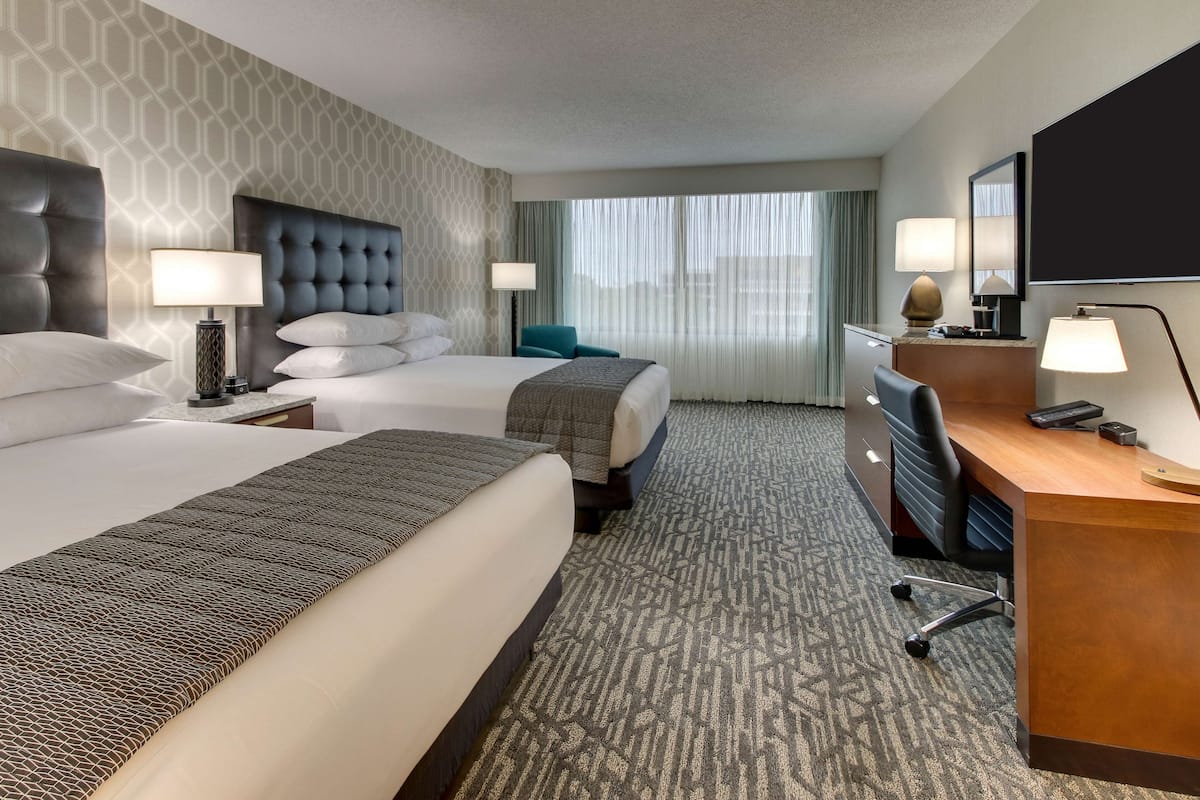 A hotel room with two beds, a desk, a chair, a TV, and a large window with curtains.