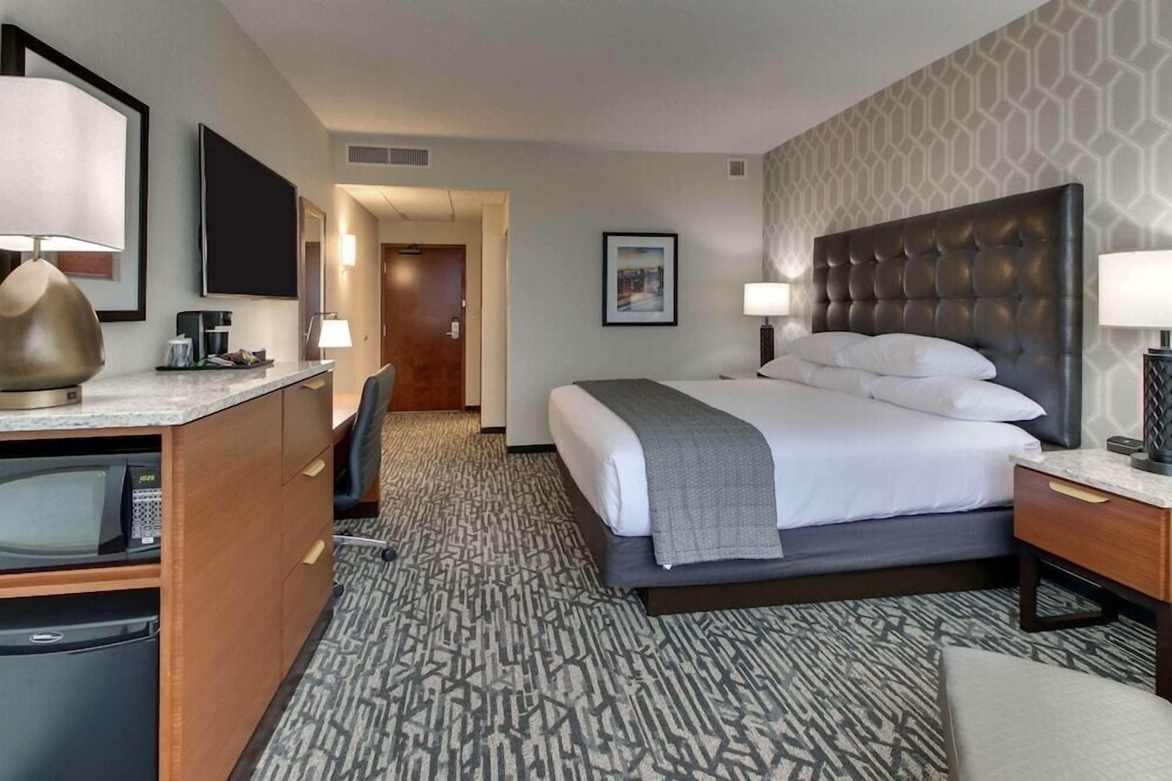 A hotel room with a bed, desk, chair, TV, and a patterned carpet.