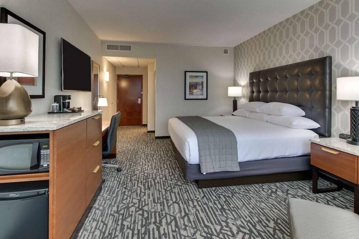 A hotel room with a bed, desk, chair, TV, and a patterned carpet.