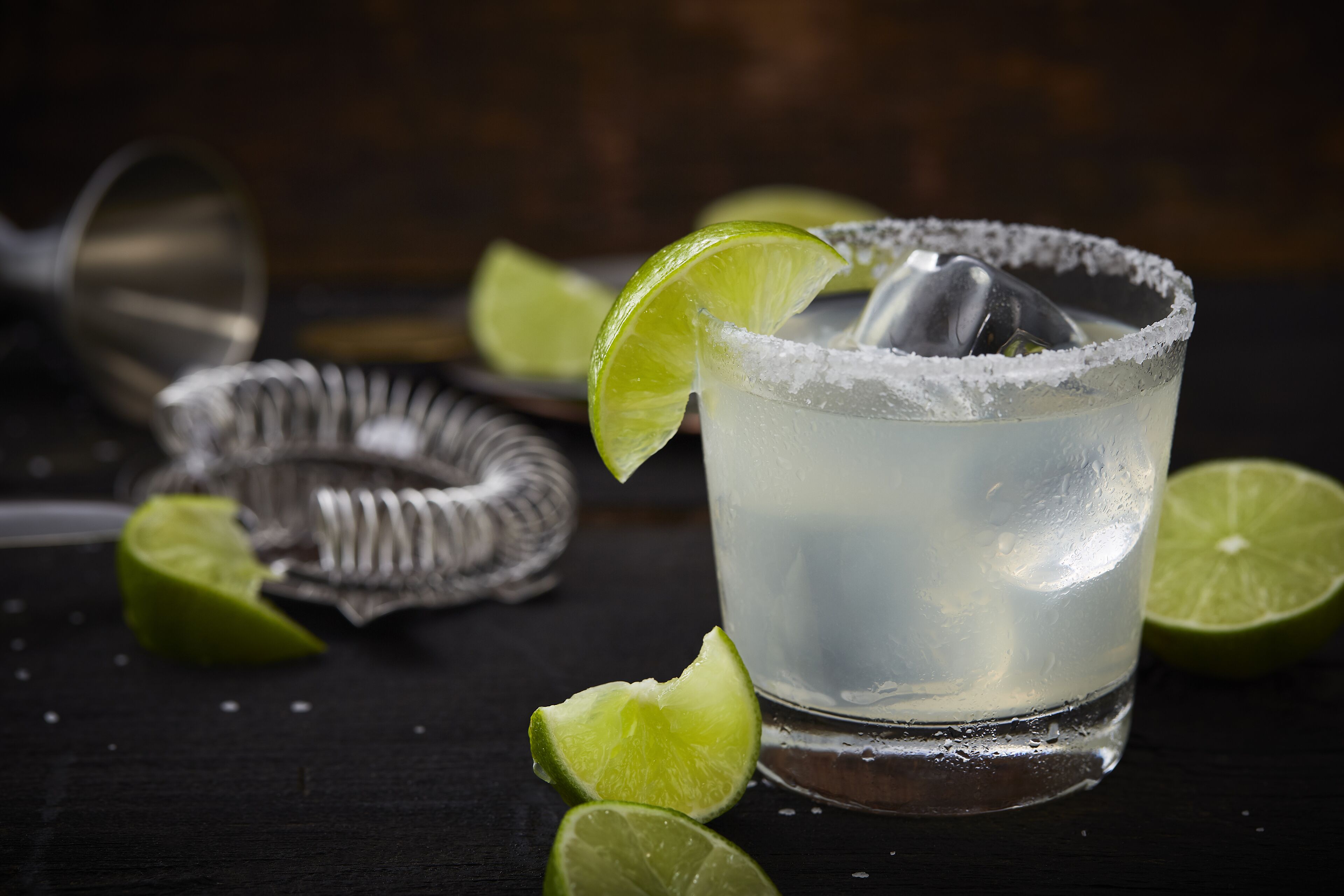A glass of a clear beverage with a lime garnish and salt rim, accompanied by a lime wedge and a metal jigger on a dark surface.