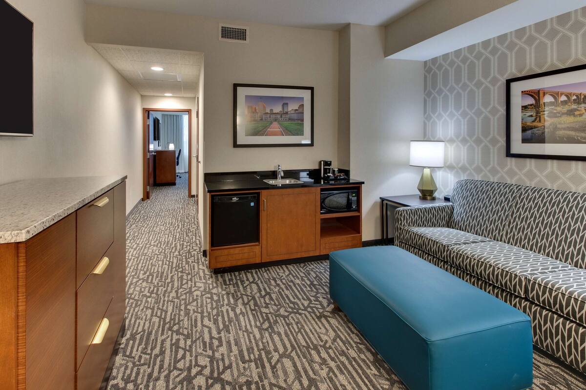 A hotel room with a kitchenette, a sofa, a bench, and a framed picture on the wall.