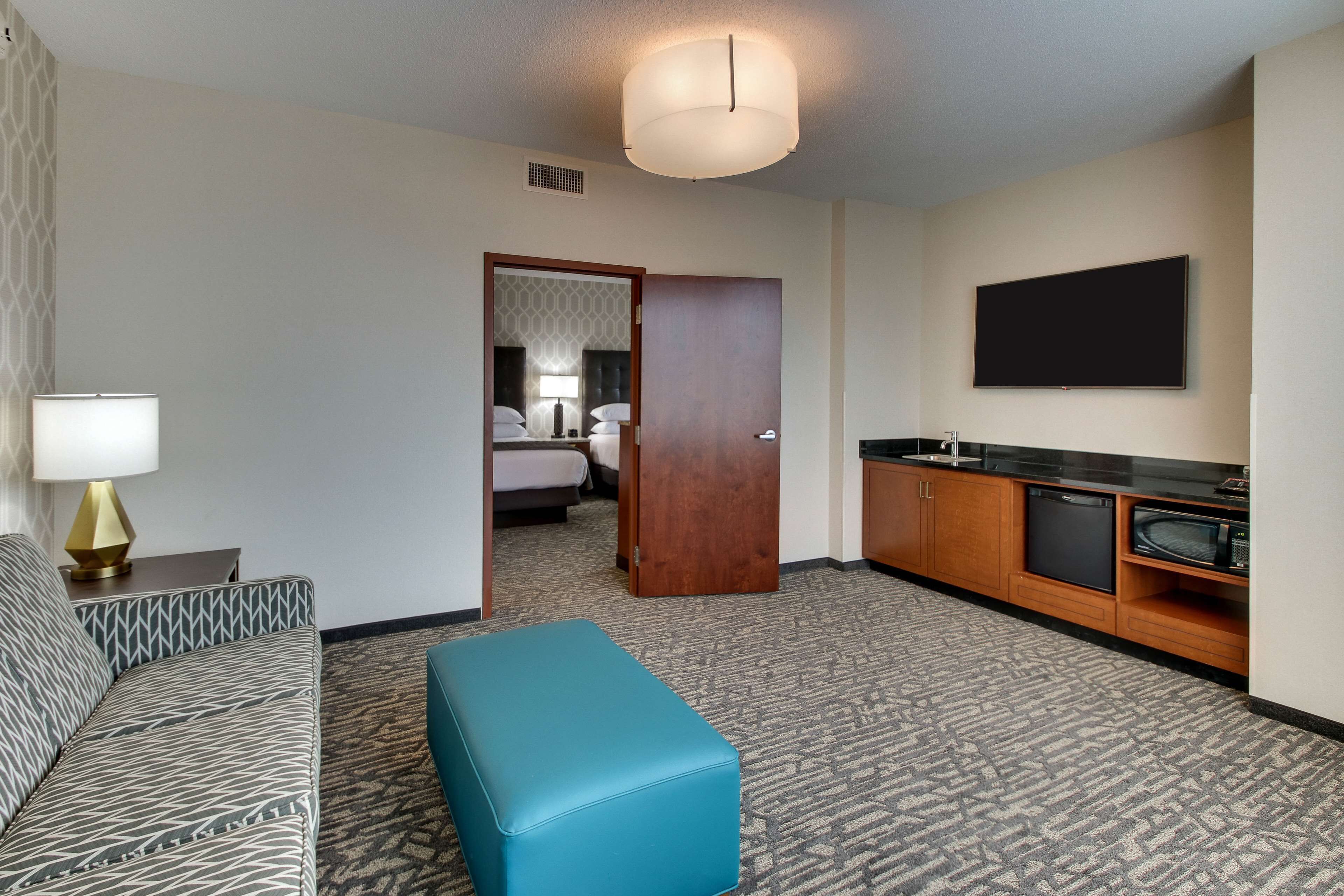 A hotel room with a patterned sofa, a teal ottoman, a flat-screen TV, a kitchenette, and a door leading to a bedroom.